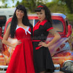 Two women in vintage-style outfits standing next to a red car.